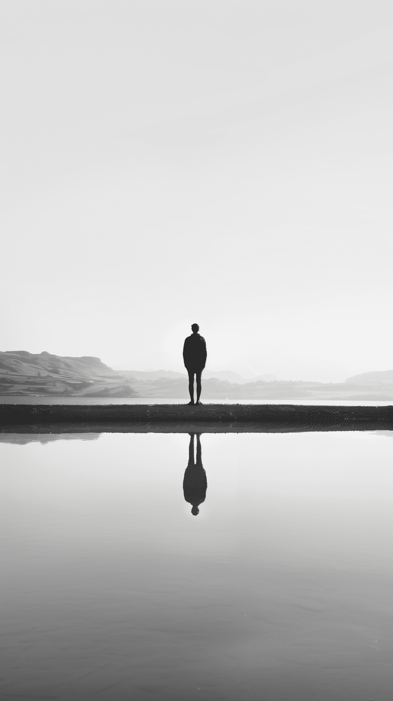 Lone figure standing at the edge of a still water body, image perfectly reflected below, almost symmetrical visual, surrounded by a vast, empty space that evokes a sense of isolation and introspection, all rendered in muted monochrome tones --chaos 1. 2 --ar 9:16 --style raw --v 6