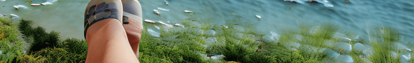 Photograph of a beautiful european woman, long ash-white hair, freckles, light-blue eyes, sitting on the edge of a dock dipping her feet into the ocean, loose tank top, shorts, warm summer day, sun shining high in the sky, sunlight reflecting off the calm water